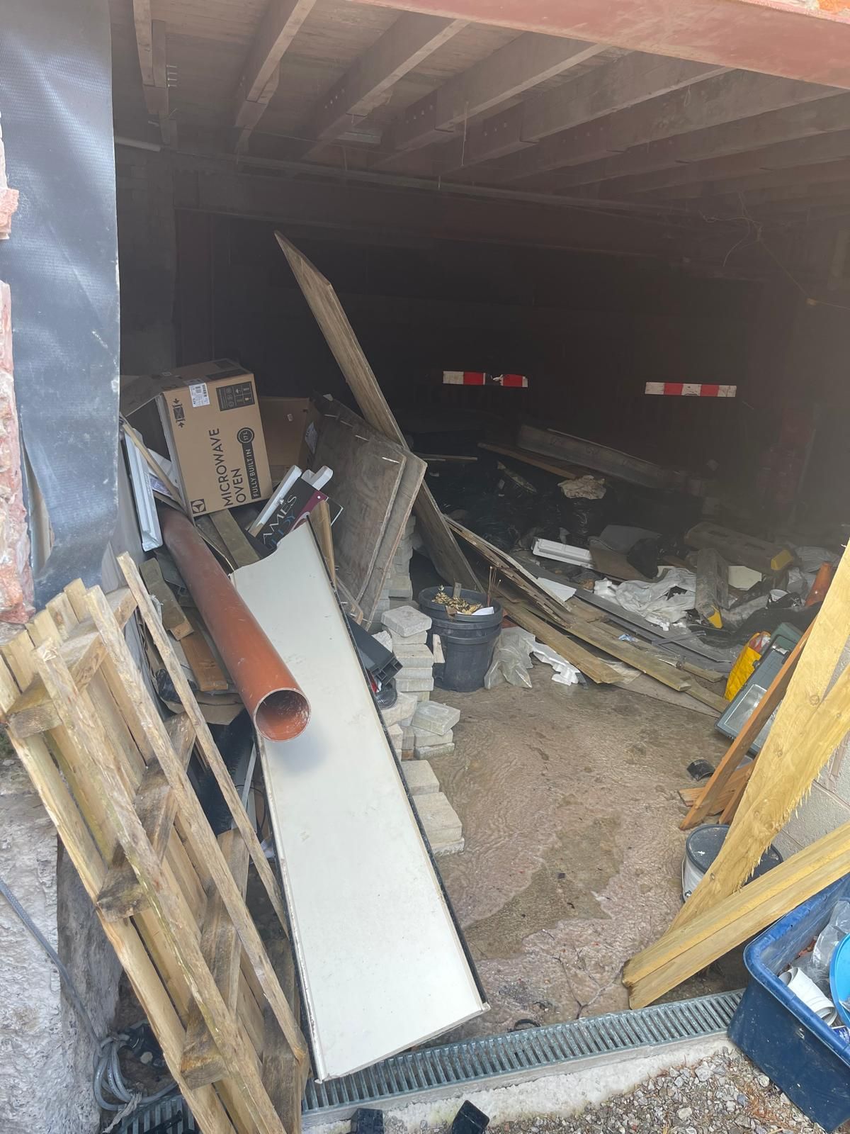Debris piled at a basement opening, including a microwave oven box, stacked paving setts, timber and an orange soil pipe, with a drainage channel in the foreground.