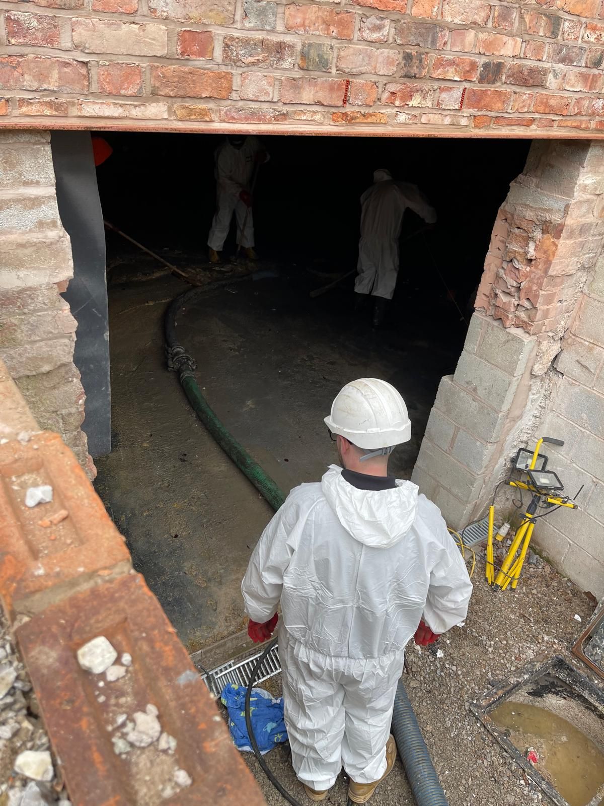 An operative in white protective coveralls and a hard hat at a basement access opening, with two more coveralled operatives clearing the dark, flooded basement behind a suction hose and task lights.