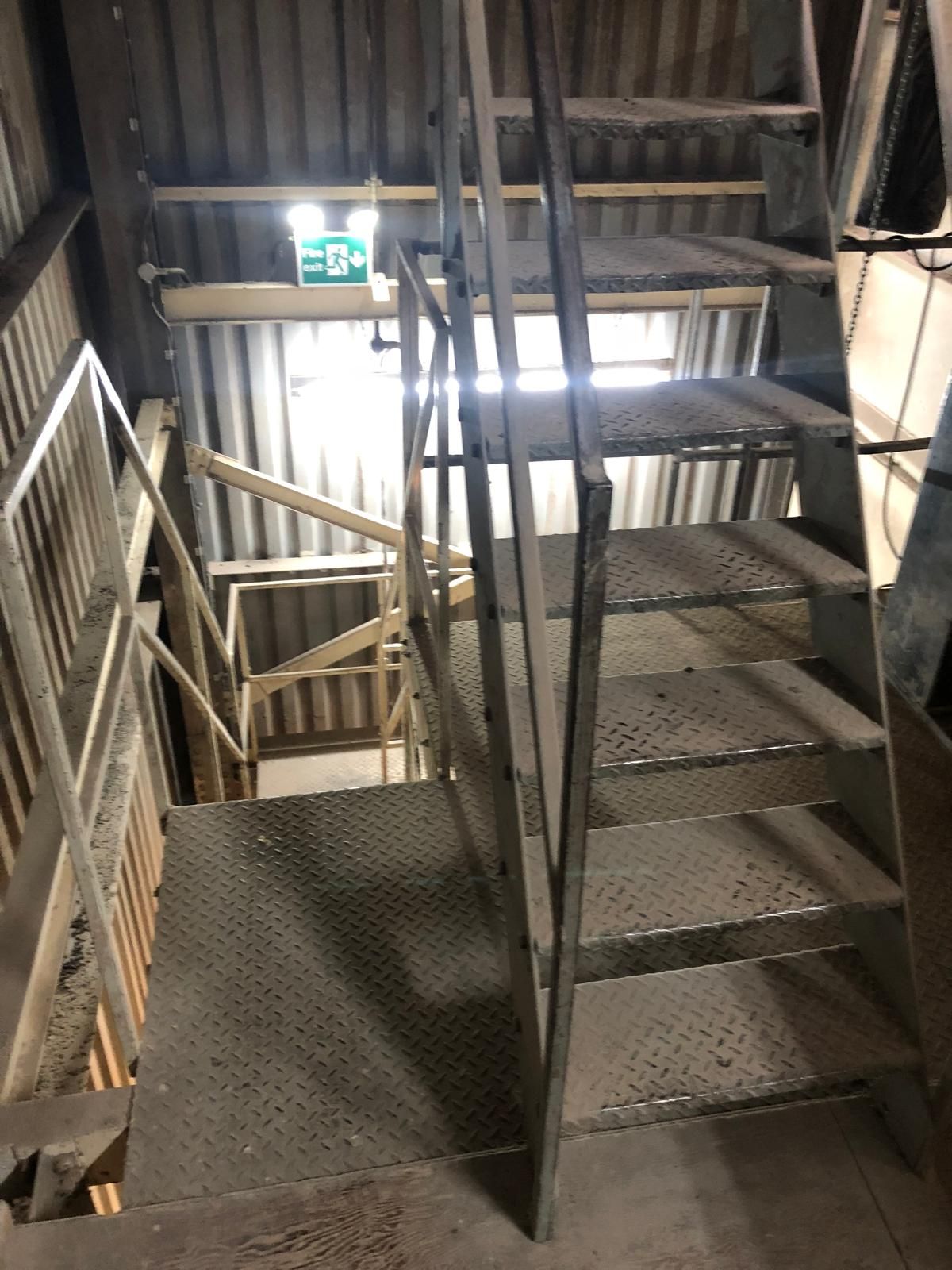 A galvanised steel access staircase with chequer-plate treads inside a corrugated-clad feed mill, lit by a fire exit sign.