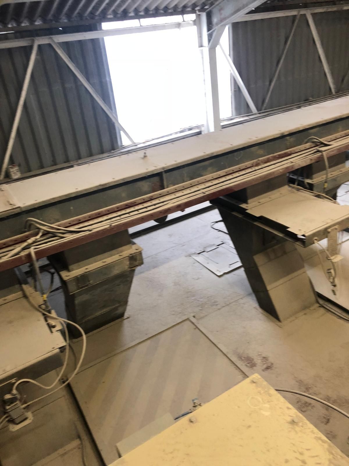 A long enclosed chain conveyor casing running the length of a feed mill on concrete plinths, above a dust-covered floor set with access hatches.