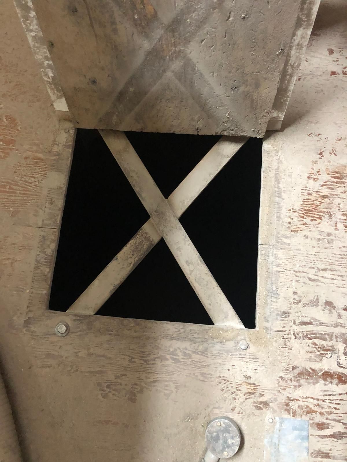 An open square access hatch in a dust-covered mill floor, with a diagonal steel cross-brace spanning the dark void of the silo below.