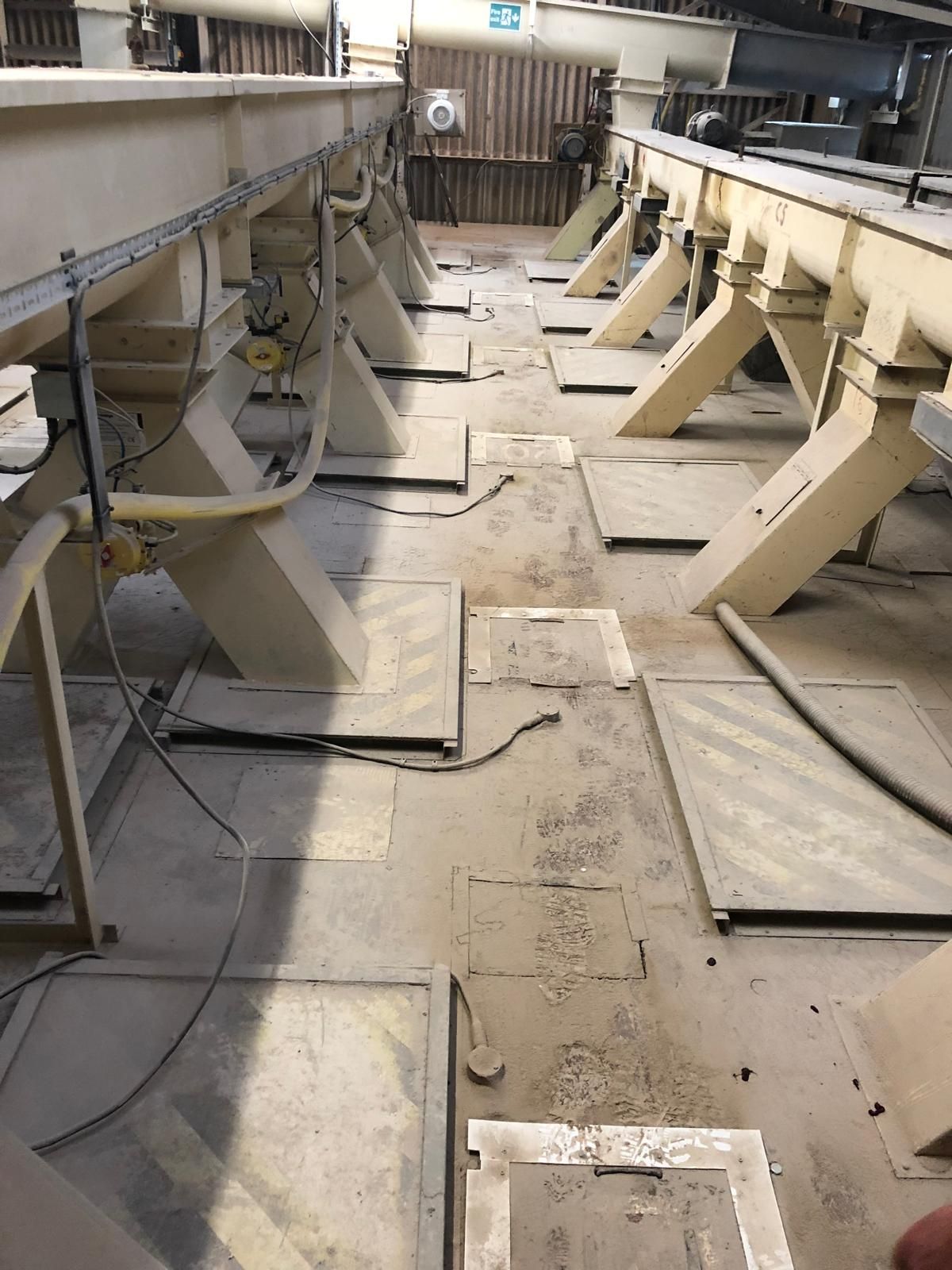 A row of beige feeder machines on steel legs standing over a mill floor covered in square metal silo inspection hatches and hazard-striped covers, under heavy dust.