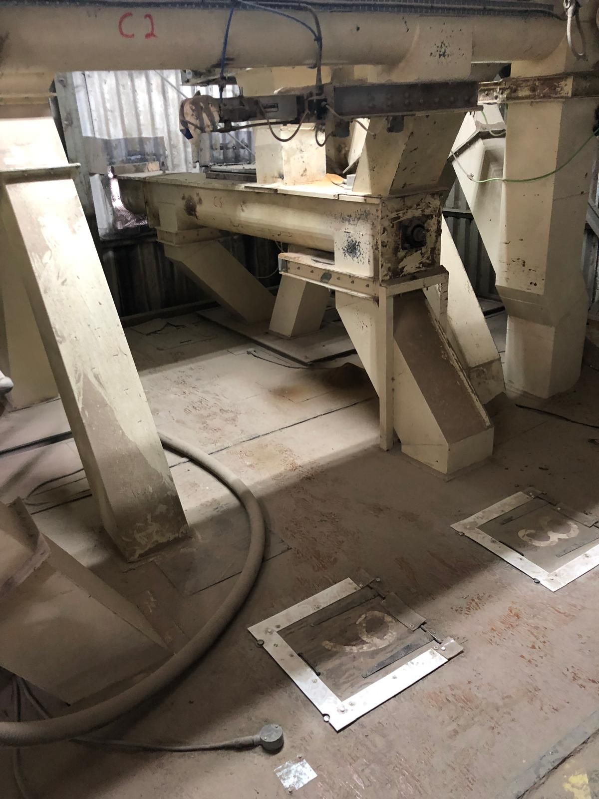The underside of a feed mill, showing a beige vibrating feeder unit on steel legs above a dust-covered floor set with square silo inspection hatches, with a flexible suction hose running across it.