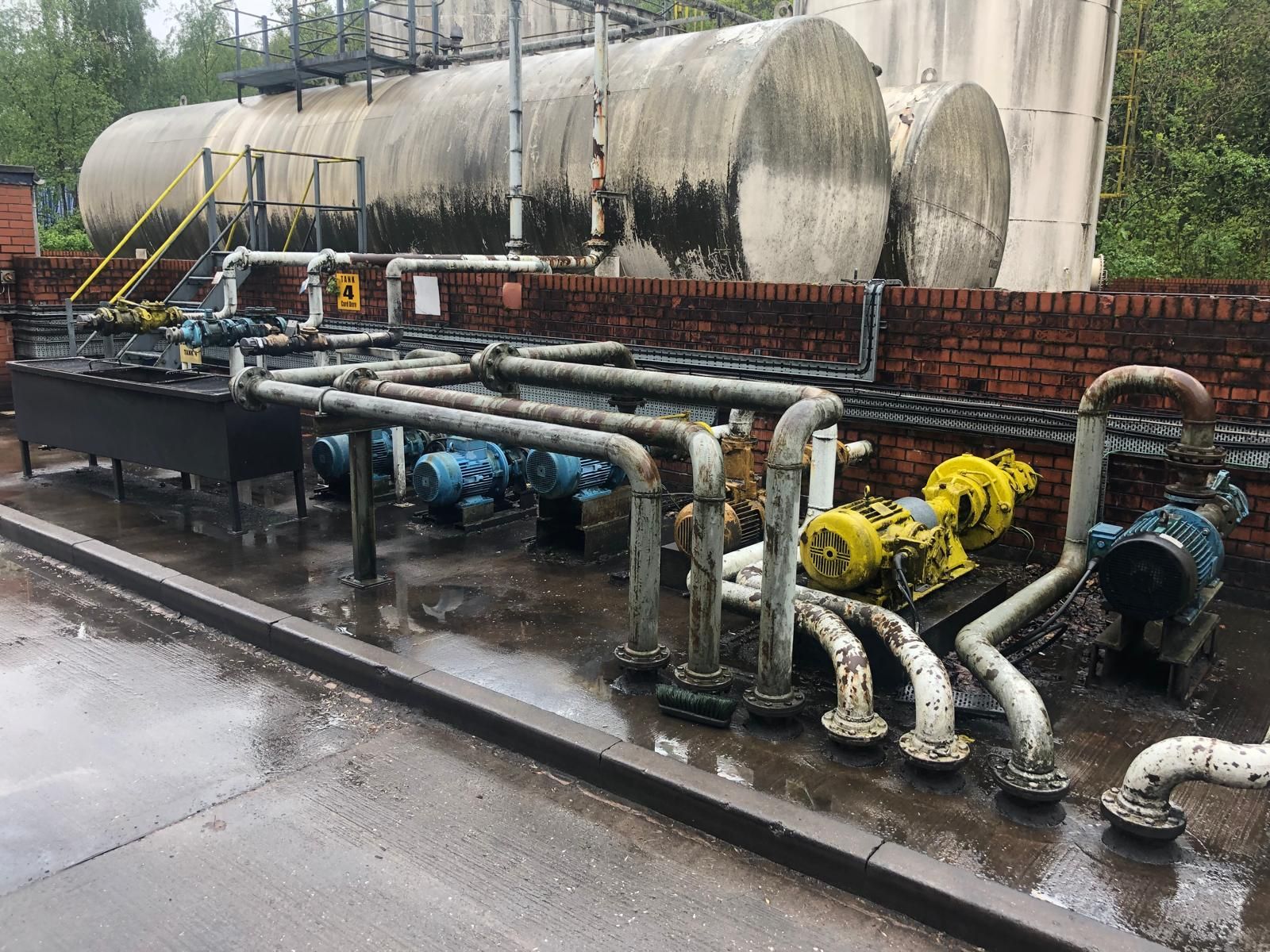 Blue and yellow fuel transfer pump motors and galvanised pipework inside the bunded compound, with horizontal storage tanks behind.