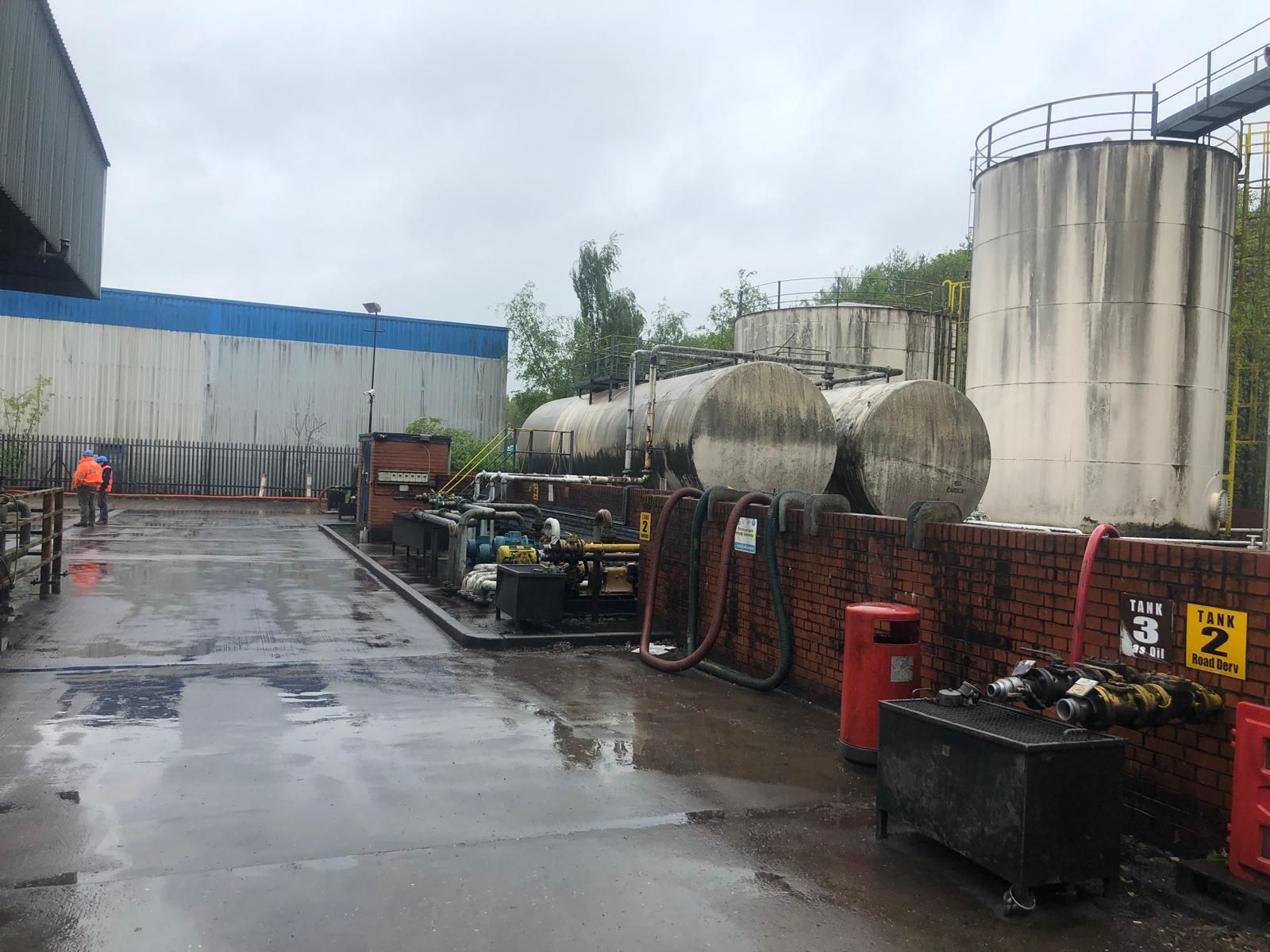 Horizontal and vertical fuel storage tanks behind a brick bund wall signed Tank 3 Gas Oil and Tank 2 Road Derv, with two operatives in orange hi-vis on site.