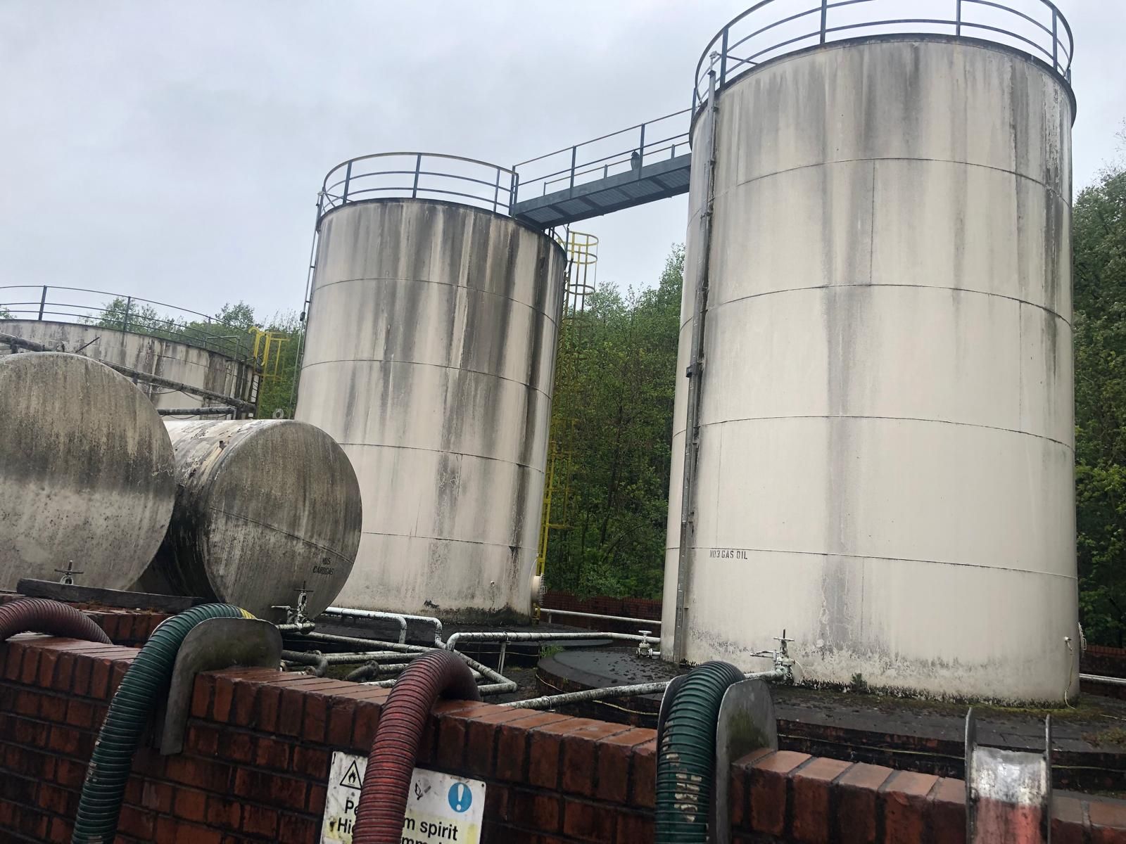 Two tall vertical fuel storage tanks joined by a walkway bridge, labelled No3 Gas Oil, with red and green suction hoses draped over the brick bund.