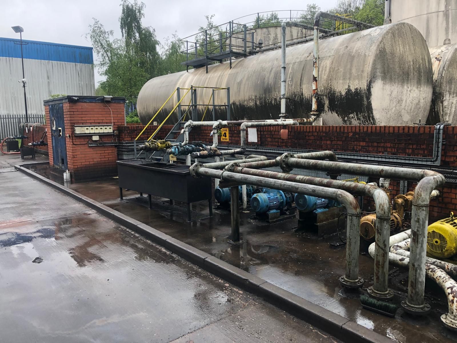 A horizontal bulk fuel tank behind a brick bund wall, with a row of transfer pump motors and galvanised pipework in the foreground at a fuel storage depot.