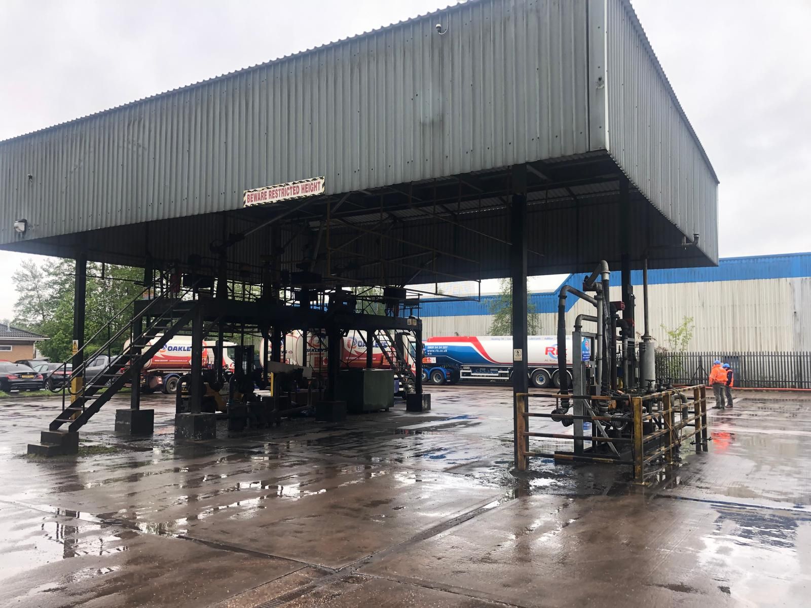 A steel loading canopy marked Beware Restricted Height, with road fuel tankers parked beneath it and two operatives in hi-vis on the wet yard.