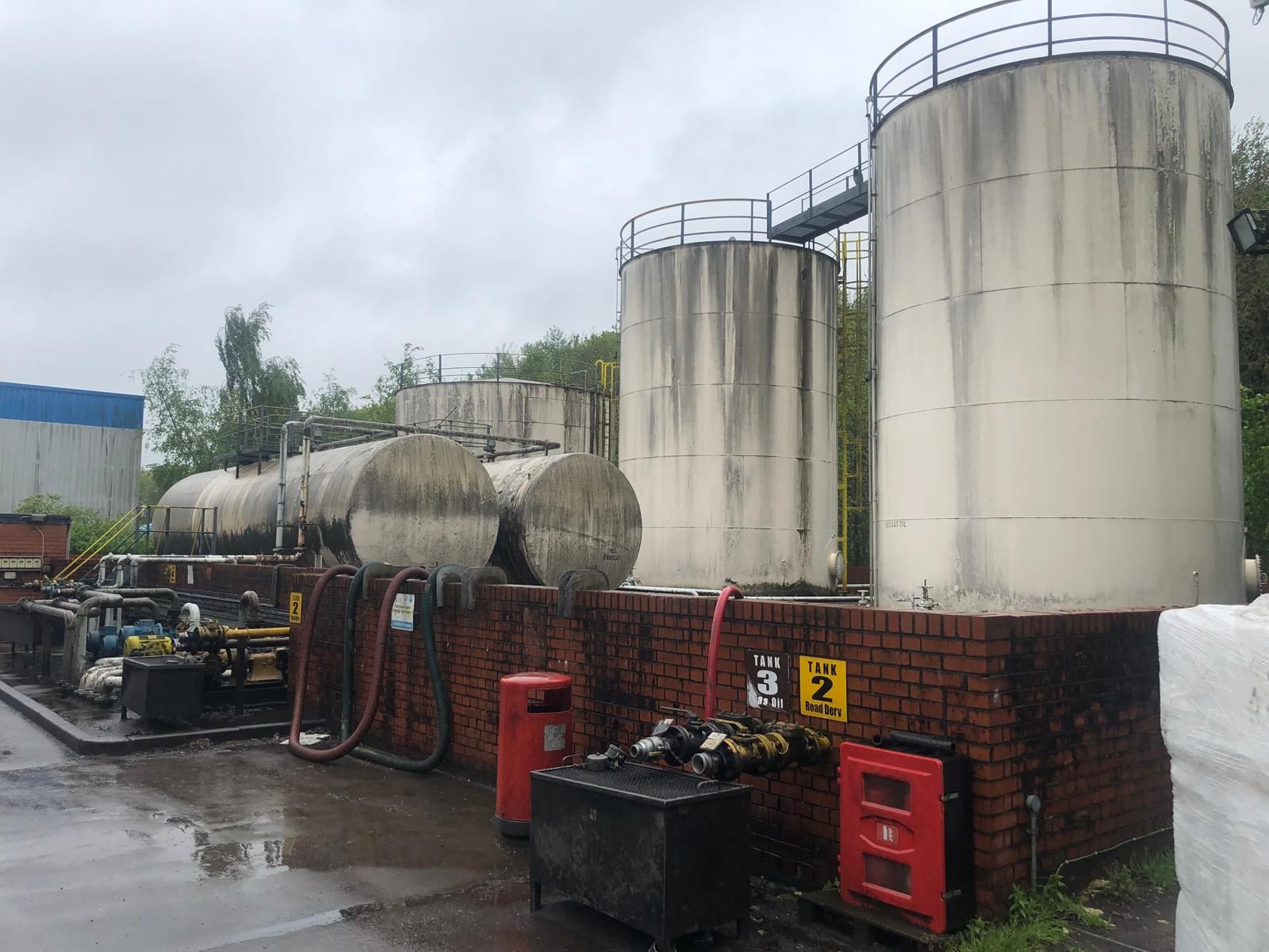 Two large vertical bulk fuel storage tanks and horizontal tanks behind a brick bund, with hose couplings and a fire point on the wall.