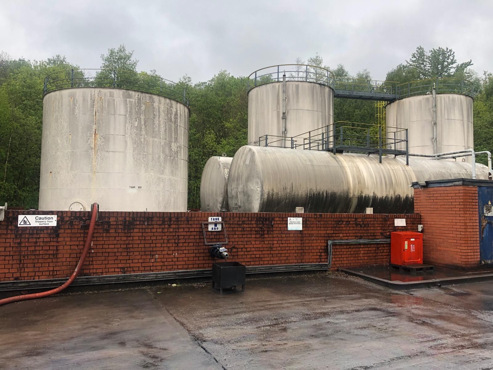 A wide view of the fuel tank farm with vertical and horizontal tanks, a Tank RBO sign and warning notices for slippery floor and highly flammable petroleum spirit.