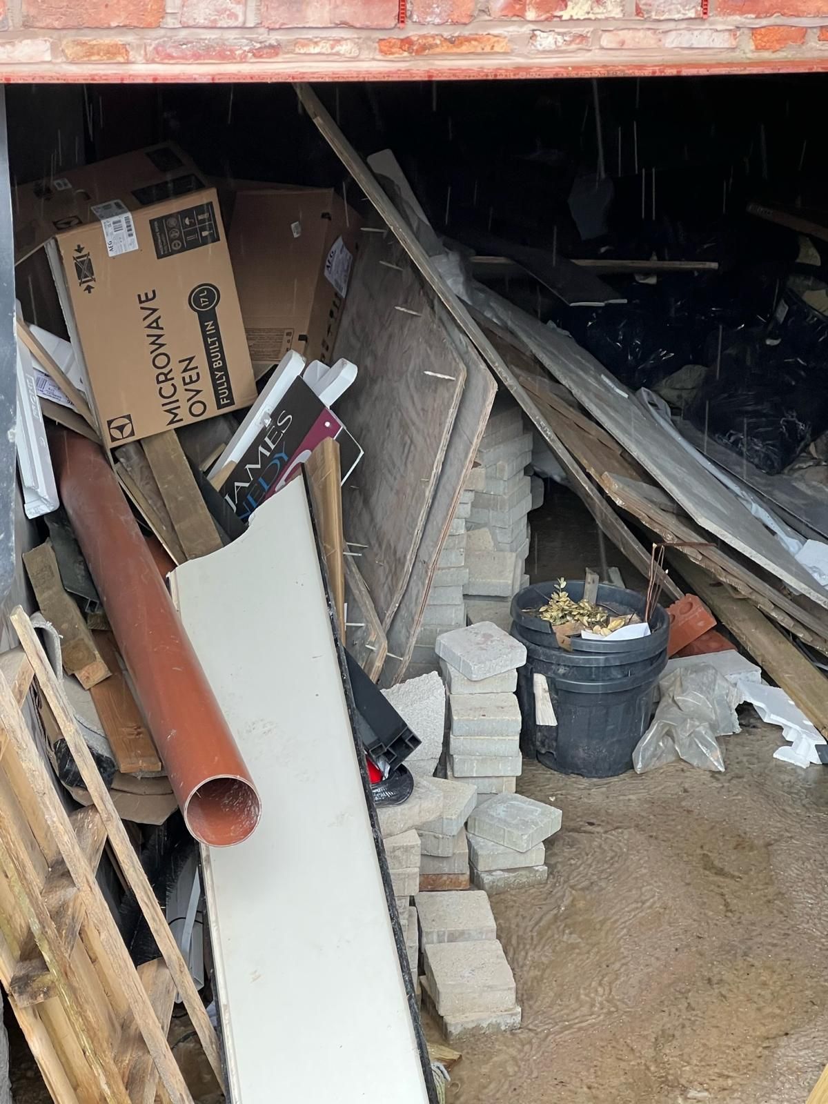 Bulky waste packed beneath a building — a large appliance box, timber, plastic pipe, paving setts and general packaging — being cleared during a site closure.