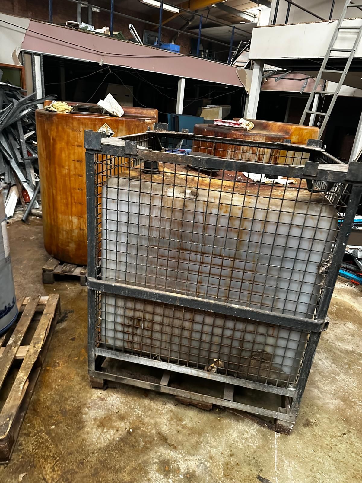 A caged intermediate bulk container beside large graduated storage tanks in a unit being cleared, with a stripped mezzanine and scrap metal behind.
