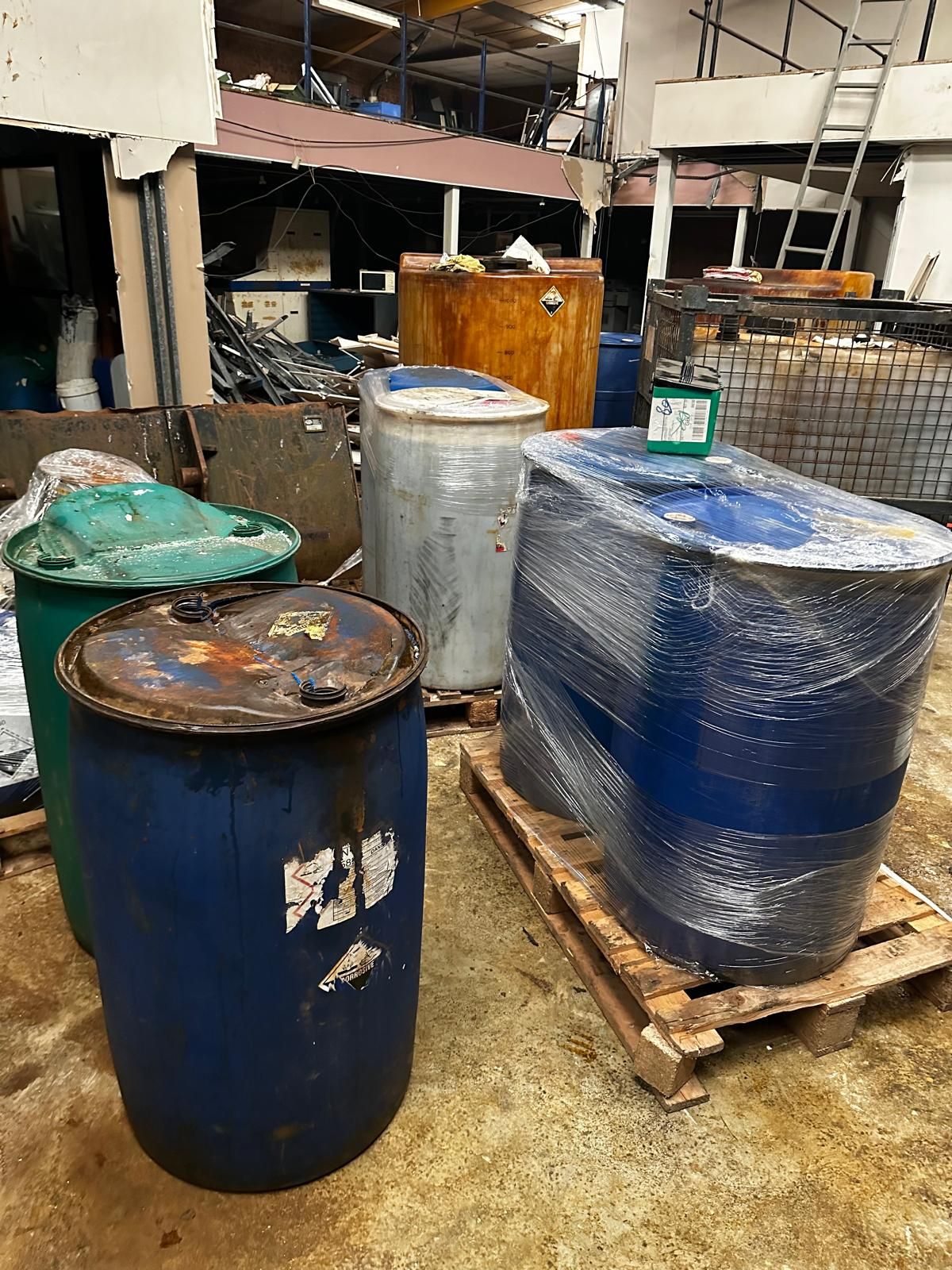 Steel and plastic drums and a wrapped bulk container standing in a vacated industrial unit, with scrap metal and a stripped mezzanine behind, ready for removal.