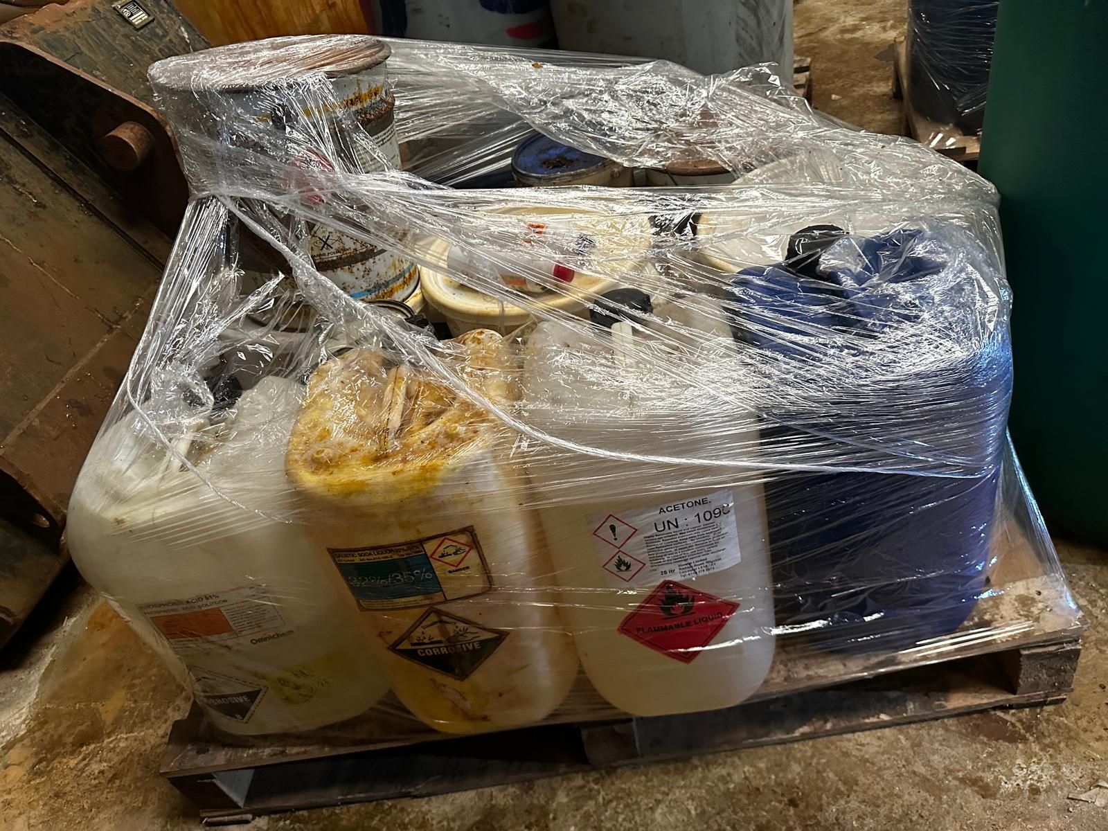 A wrapped pallet of jerrycans of caustic soda, acetone and phosphoric acid carrying corrosive and flammable hazard labels, segregated for compliant disposal during a site closure clearance.