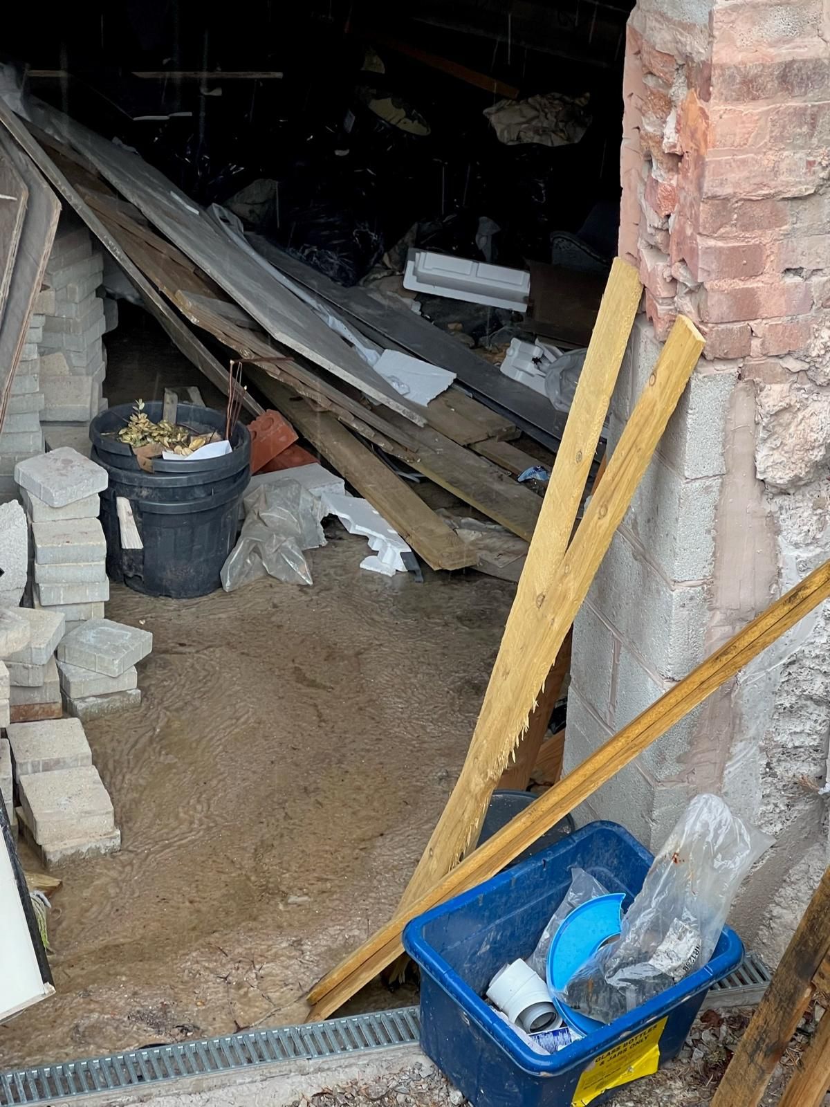 A low storage void beneath a building being cleared of stacked paving blocks, timber offcuts, polystyrene and mixed household waste, with a brick pier opened up alongside.