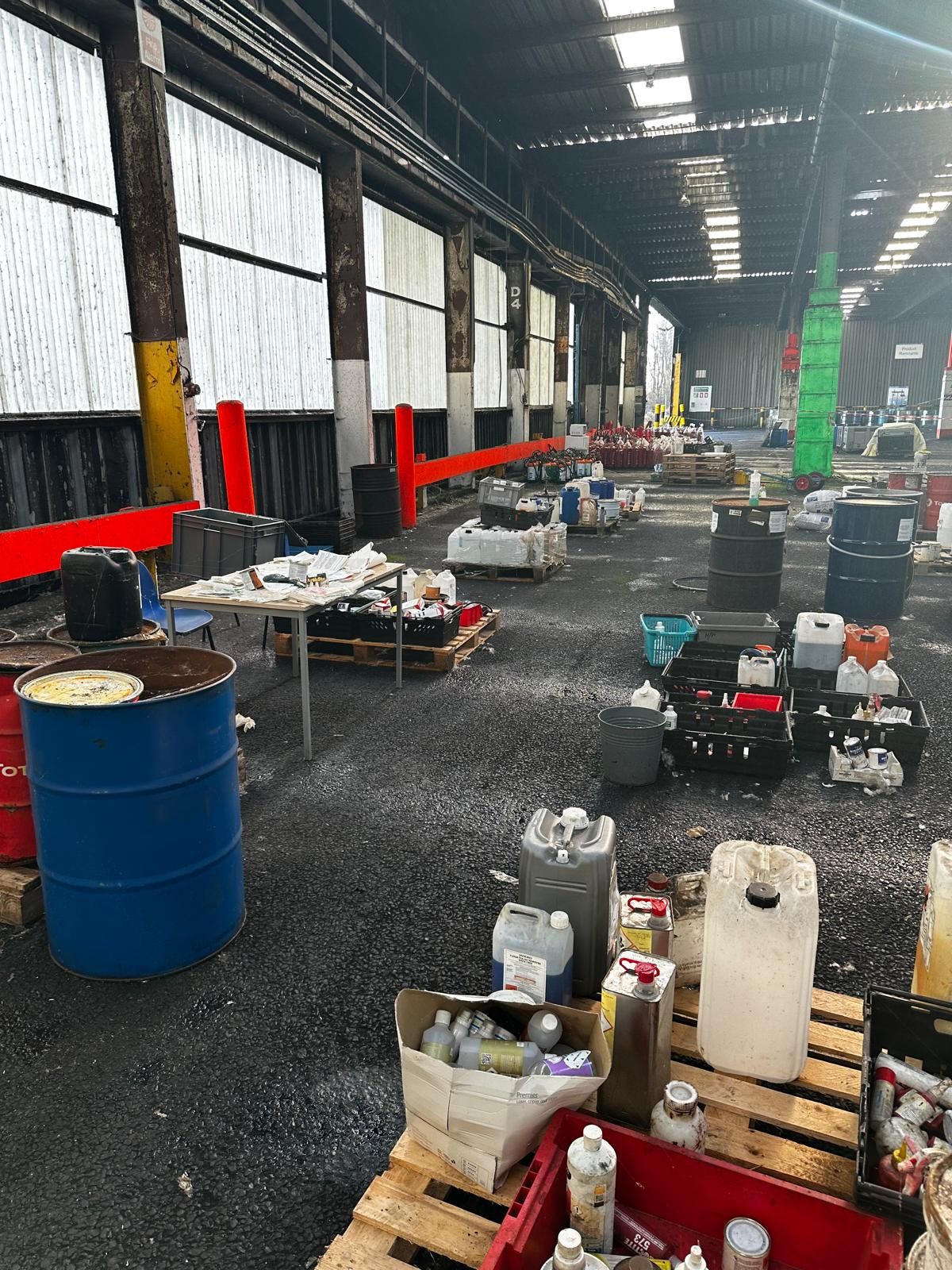 The interior of a large industrial warehouse being cleared during a site closure, with drums, pallets of chemical containers and crates of part-used products segregated across the floor.