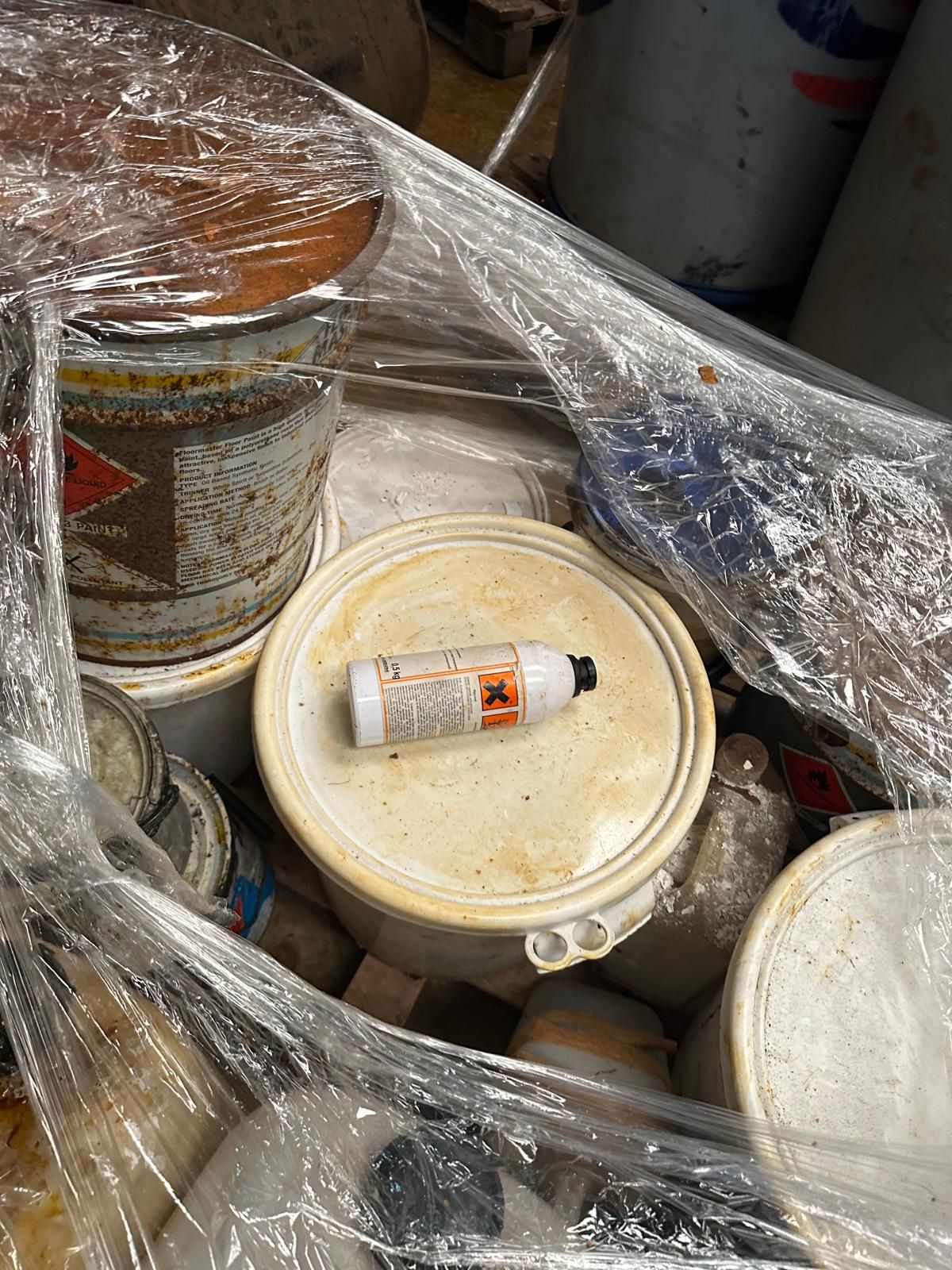 A shrink-wrapped pallet of old paint and coating tins, one marked with a harmful-substance hazard symbol, gathered for classification during a site closure clearance.