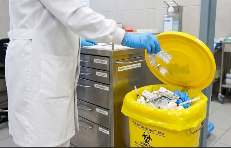 A healthcare worker in gloves placing medicines into a yellow clinical waste container in a dispensary.